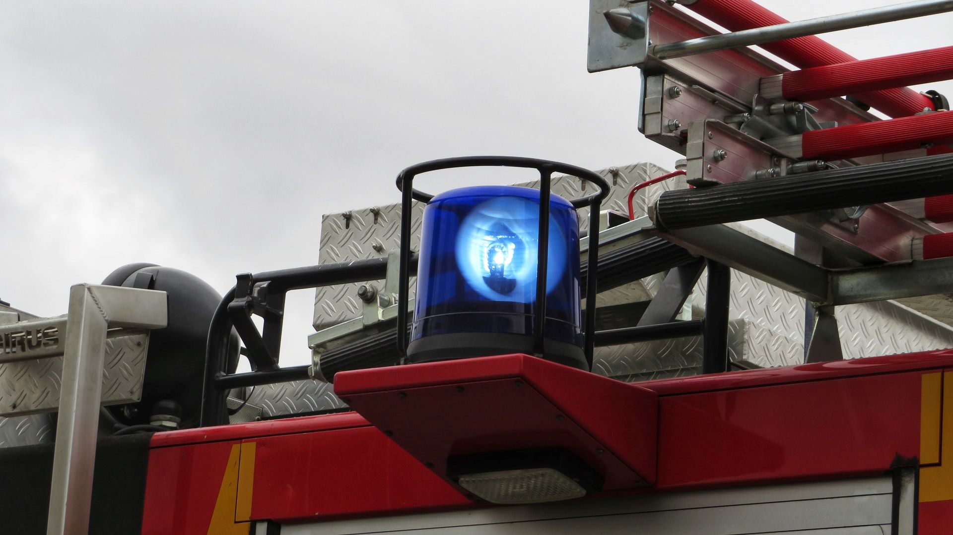 Das Bild zeigt ein Feuerwehrfahrzeug, auf dem eine blaue Rundumleuchte montiert ist. Die Leuchte befindet sich auf einem roten Aufbau des Fahrzeugs, umgeben von metallischen Komponenten und Ausrüstungen, die typisch für Feuerwehrfahrzeuge sind. Die blaue Leuchte ist eingeschaltet und signalisiert die Präsenz und Einsatzbereitschaft des Fahrzeugs im Notfall. Die Anordnung und Ausstattung des Fahrzeugs deutet auf seine spezielle Nutzung im Rettungs- und Brandbekämpfungseinsatz hin.